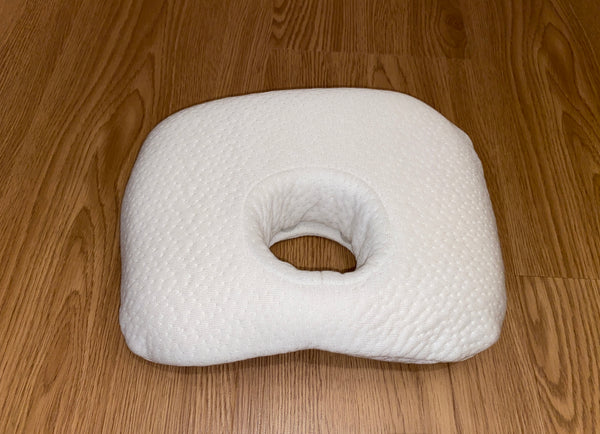 A white baby comfort pillow for head support, featuring a soft, textured cover and a central hole to help prevent flat head, shown on a light wood floor.