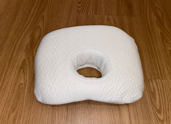 A white baby comfort pillow for head support, featuring a soft, textured cover and a central hole to help prevent flat head, shown on a light wood floor.