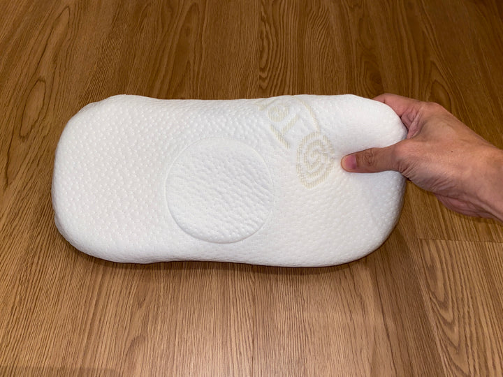 A hand squeezes a white contour baby pillow, demonstrating the soft, responsive cushion of its natural latex material for gentle head and neck support.