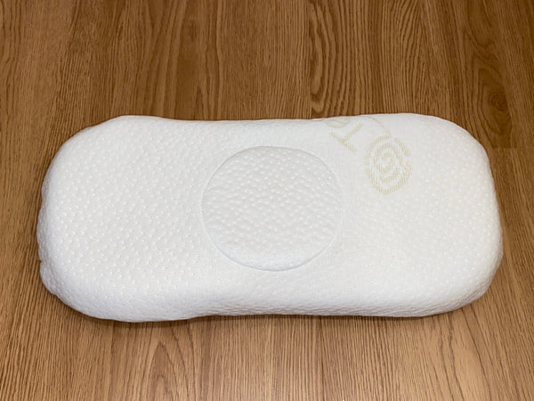 An elongated, white latex baby pillow with a central circular indentation for optimal head and neck support shown on a light wood floor.