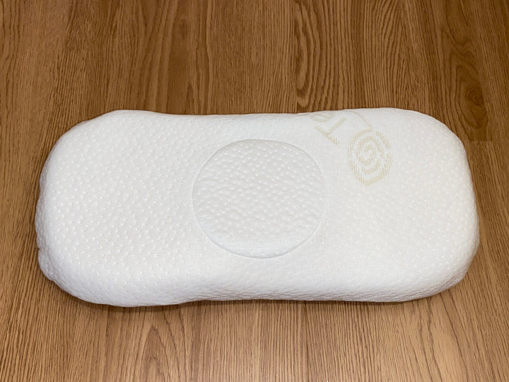An elongated, white latex baby pillow with a central circular indentation for optimal head and neck support shown on a light wood floor.
